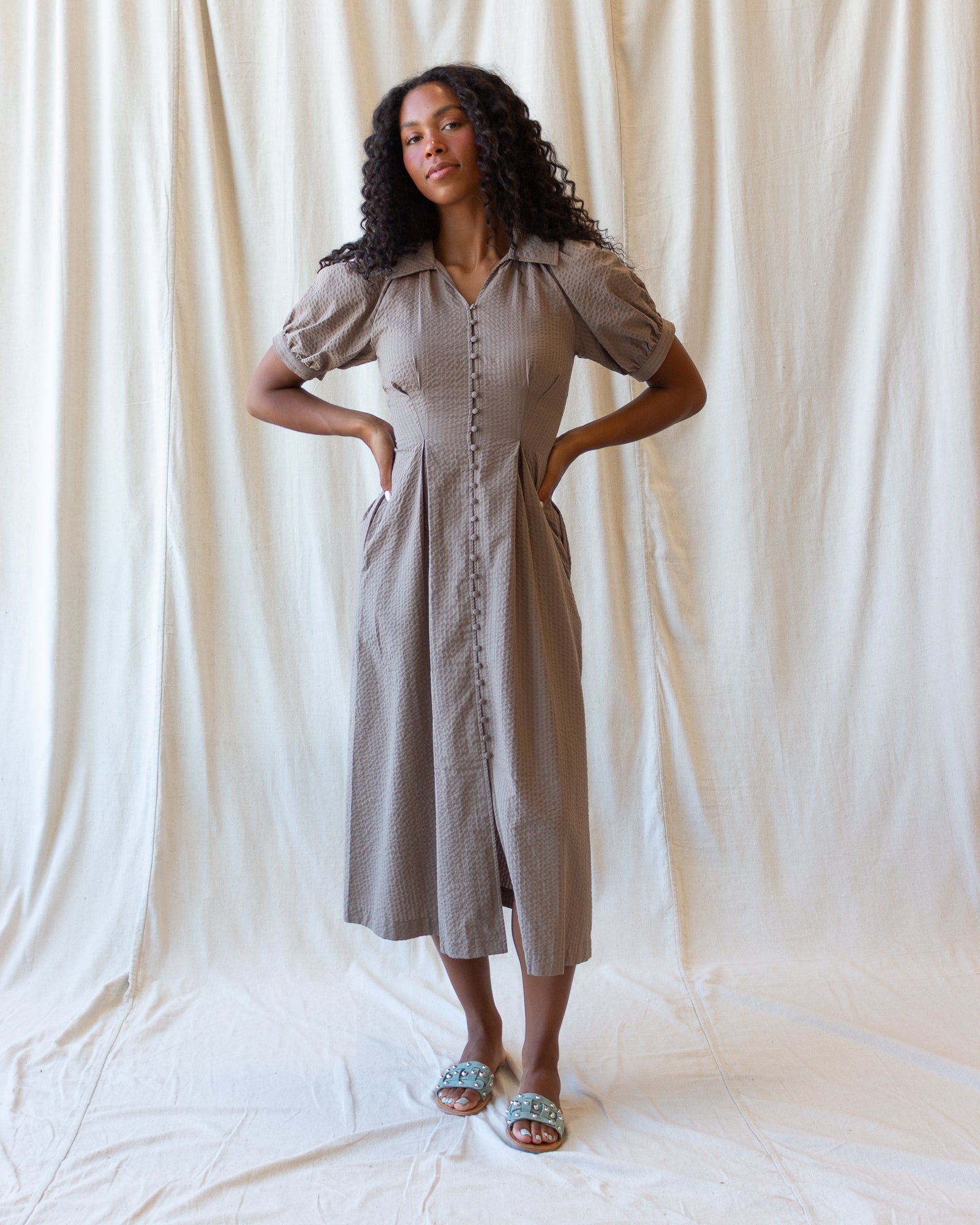 A woman modeling a taupe-colored midi dress with puff sleeves, a button-down front, and side pockets, standing against a draped cream fabric backdrop.