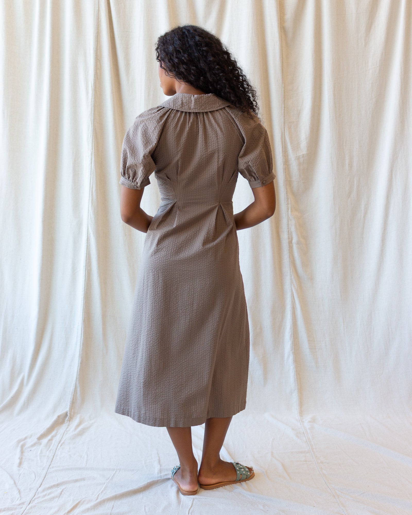 A woman stands with her back to the camera wearing a taupe brown midi dress with short puff sleeves and a gathered waist, posed against a draped cream fabric backdrop.