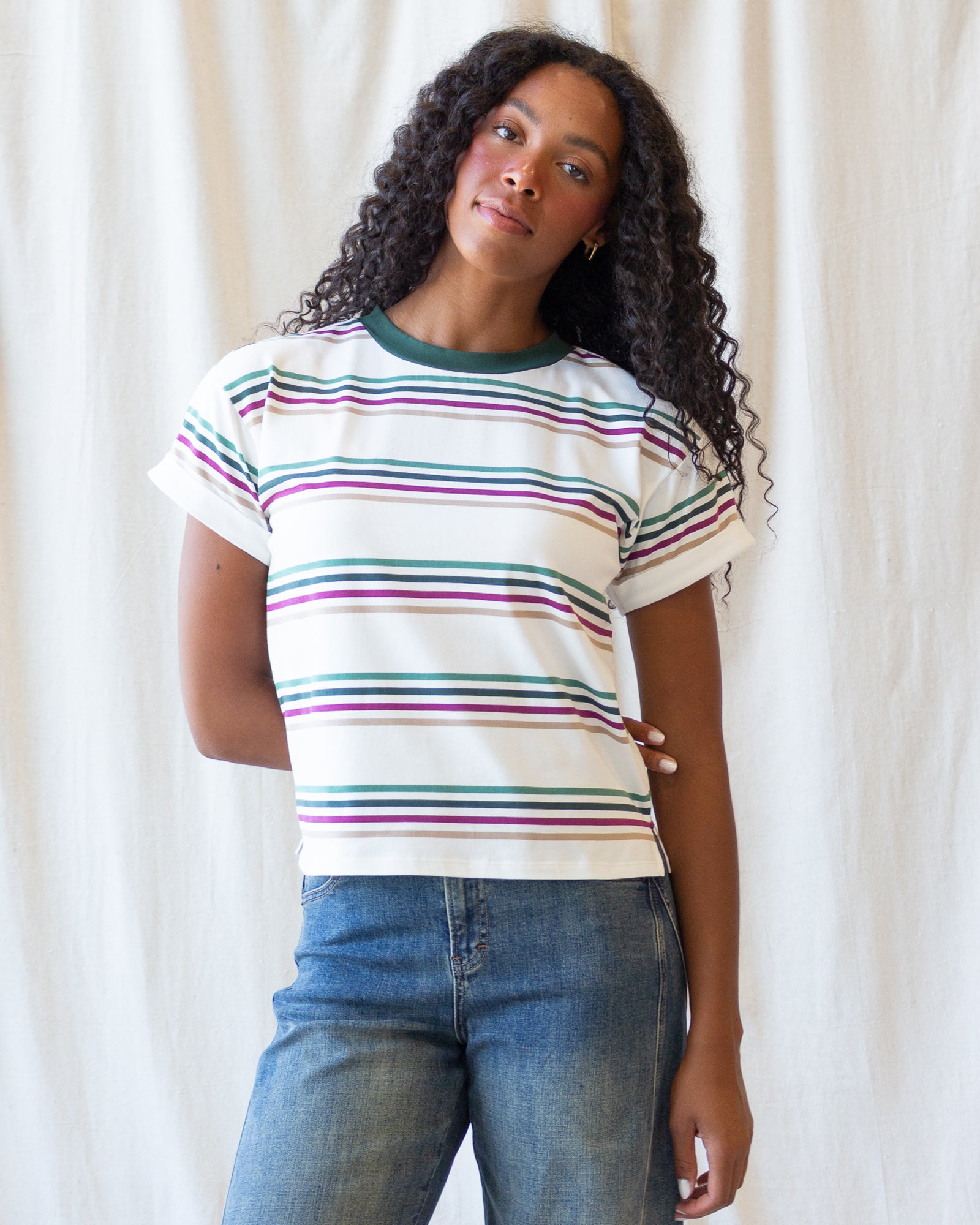 A woman with long curly hair wearing a white short-sleeve t-shirt with thin horizontal stripes in green, purple, and pink, paired with blue jeans, standing against a white fabric backdrop.