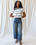 A woman stands against a draped white fabric backdrop wearing a white short-sleeve T-shirt with thin green, blue, and red horizontal stripes, blue wide-leg cropped jeans, and brown strappy sandals.