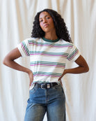 A woman with curly black hair models a short-sleeve crewneck t-shirt featuring horizontal stripes in green, purple, and pink on an off-white base. She is wearing blue jeans and a black belt, standing against a draped cream fabric backdrop.