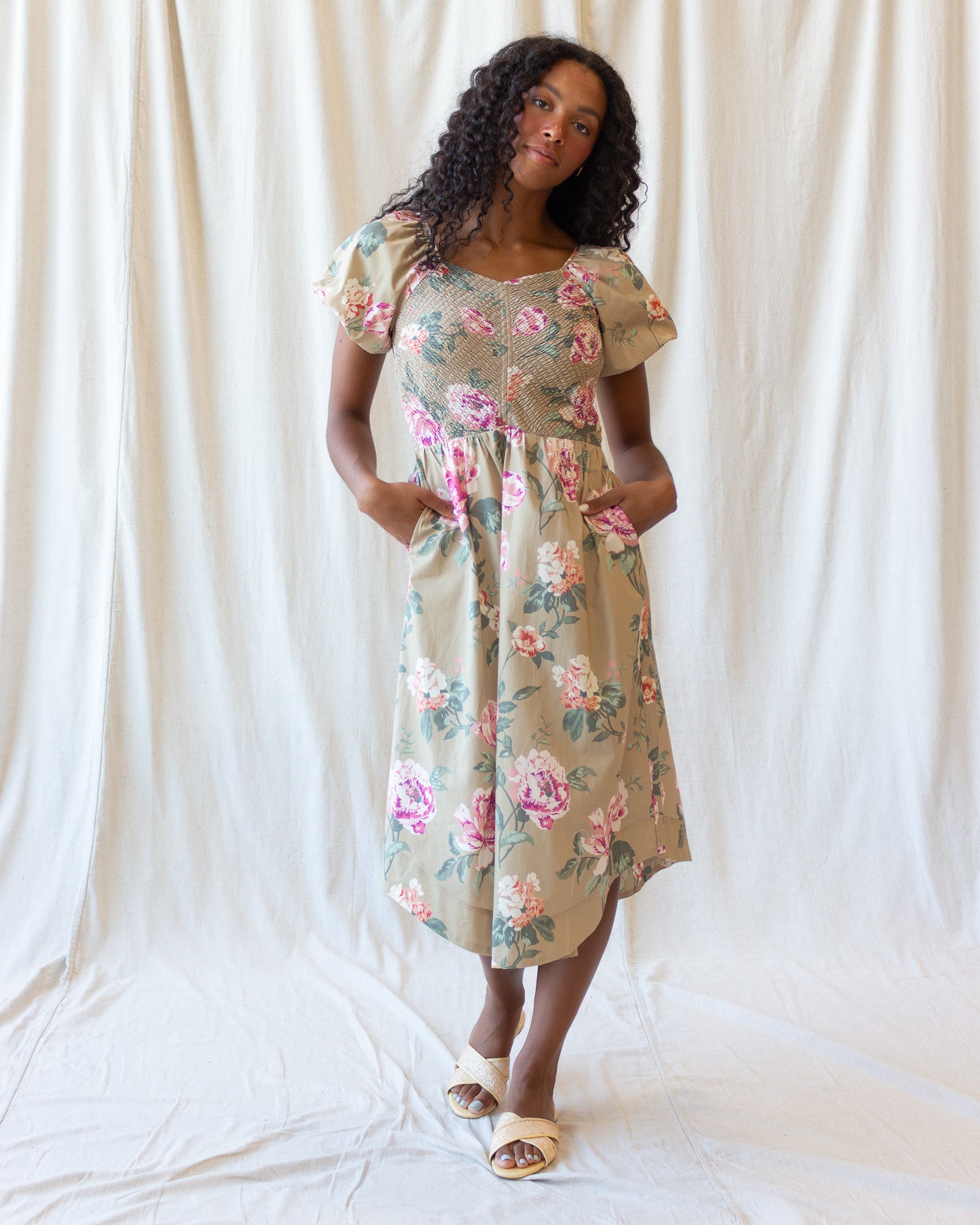 A woman modeling a short-sleeve midi dress with a beige base and a pink and green floral print, standing against a draped white fabric backdrop.