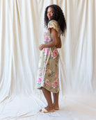 A woman modeling a knee-length floral dress with puff sleeves, standing in profile against a draped cream fabric backdrop.
