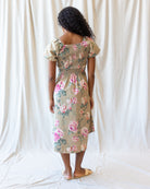 A woman viewed from behind wearing a beige midi dress with a pink and green floral print, standing against a draped white fabric backdrop.
