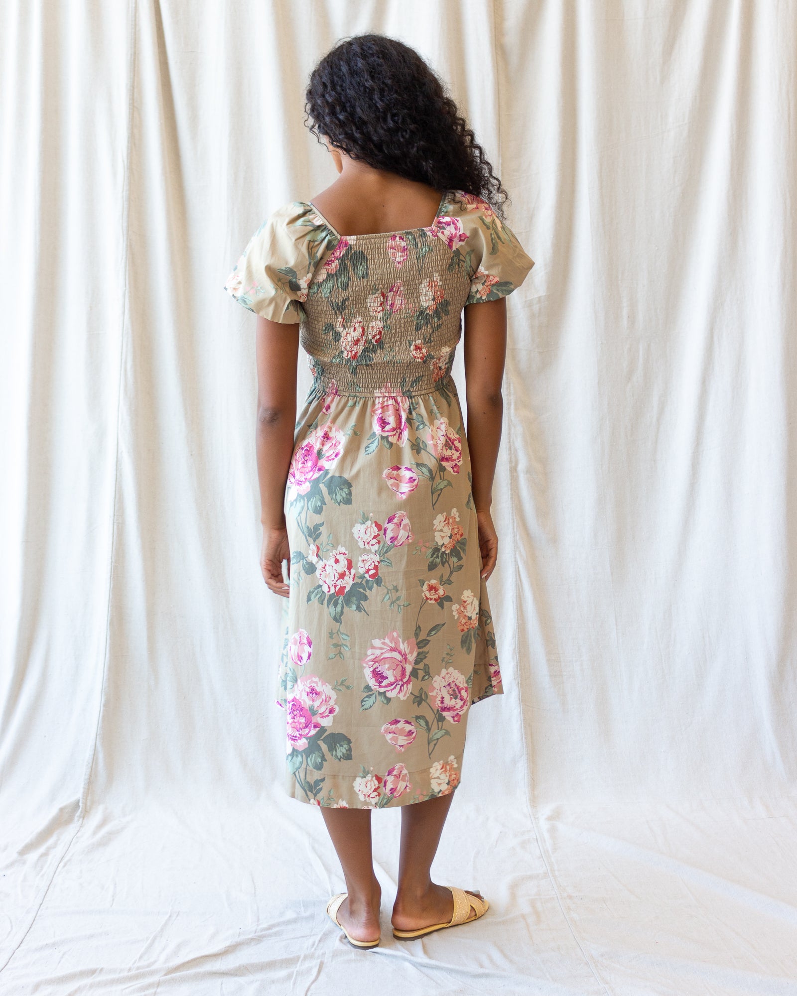 A woman viewed from behind wearing a beige midi dress with a pink and green floral print, standing against a draped white fabric backdrop.