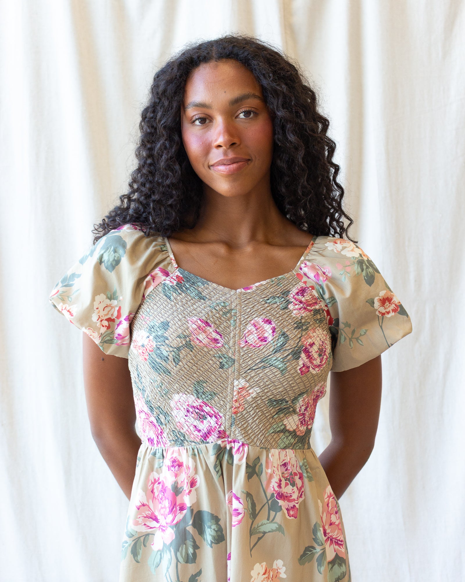 A woman with long, curly black hair wearing a beige short-sleeve dress with a pink and green floral print, standing against a white curtain background.