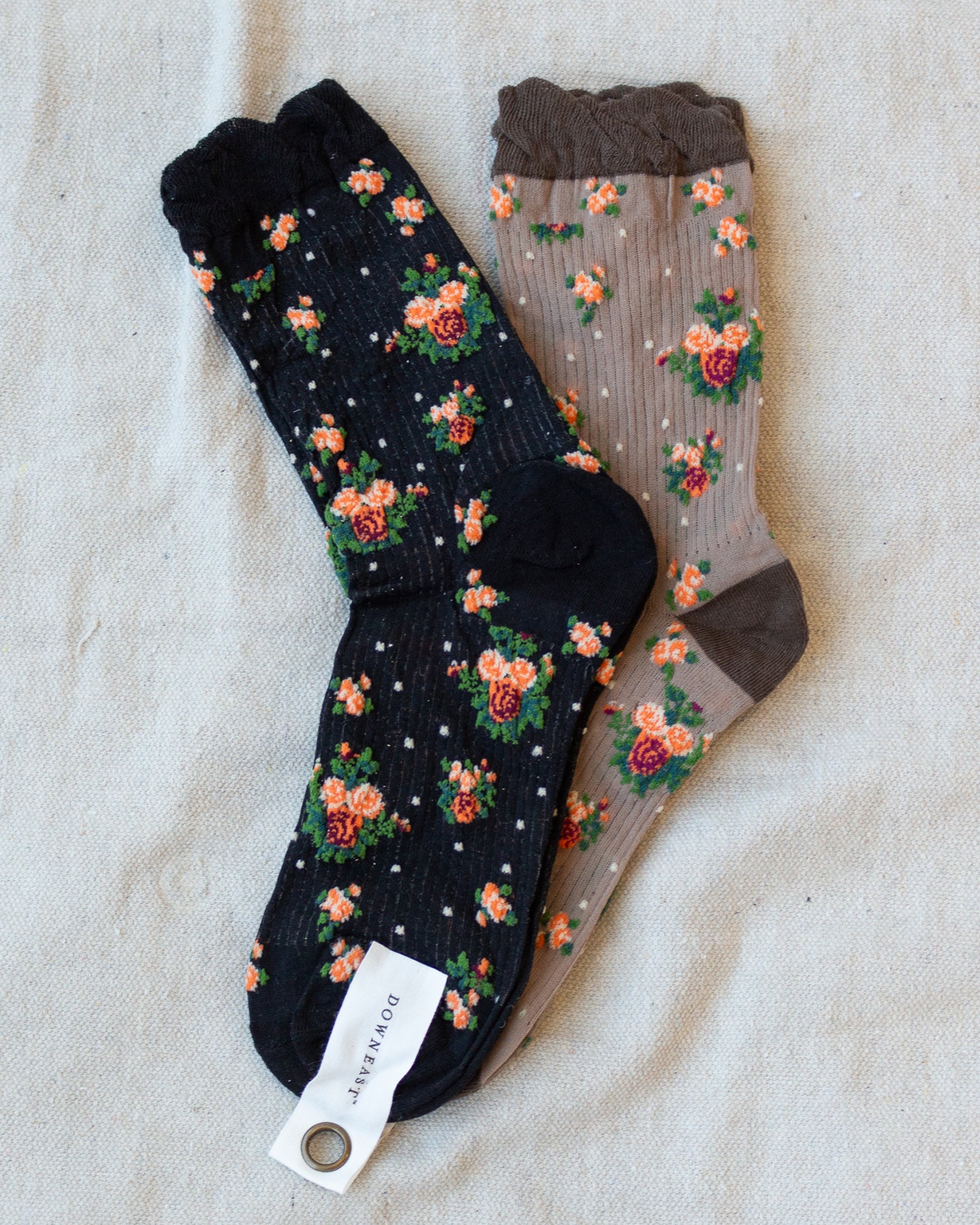 Two pairs of floral crew socks laid flat on a light beige fabric background. One pair is black with orange, pink, and green floral motifs and small white dots; the other is taupe-beige with a similar floral print and a dark brown cuff and heel.