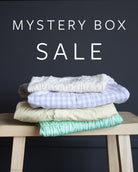 A stack of four folded items sits on a light wood stool against a dark charcoal background, with large white text reading 'MYSTERY BOX SALE' above it.