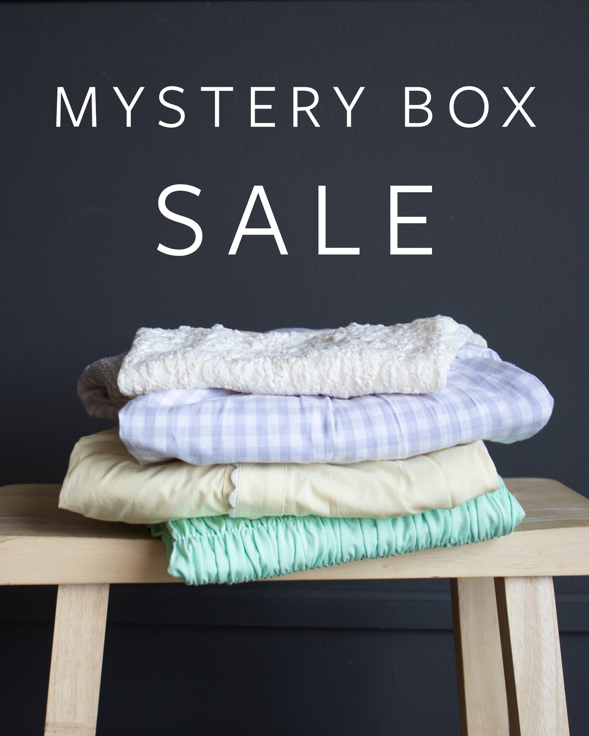 A stack of four folded items sits on a light wood stool against a dark charcoal background, with large white text reading 'MYSTERY BOX SALE' above it.