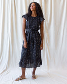 A woman modeling a dark navy midi dress with a delicate white and gold floral print, standing against a draped off-white fabric backdrop.