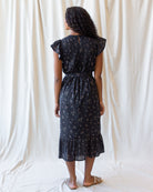 A woman stands with her back to the camera wearing a dark midi dress with a small white floral print, ruffled cap sleeves, and a ruffled hem, set against a draped white fabric backdrop.
