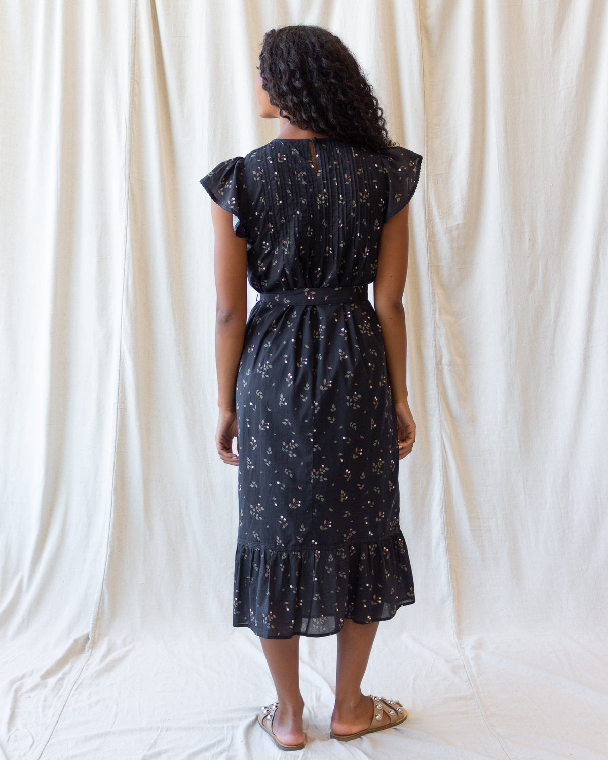 A woman stands with her back to the camera wearing a dark midi dress with a small white floral print, ruffled cap sleeves, and a ruffled hem, set against a draped white fabric backdrop.