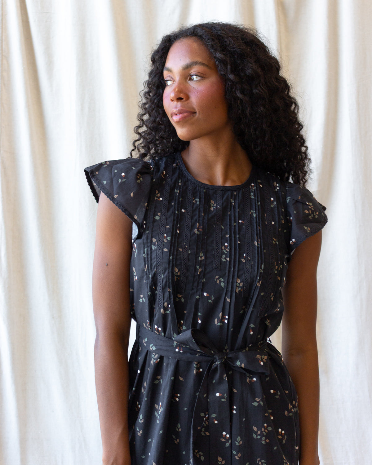 A woman with curly black hair wearing a black short-sleeve dress with a small white and gold floral print, standing against a draped cream fabric backdrop.
