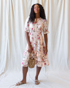 A woman stands against a draped white fabric backdrop wearing a short-sleeve cream dress with a red and pink floral print, holding a small woven bag and wearing flat sandals.