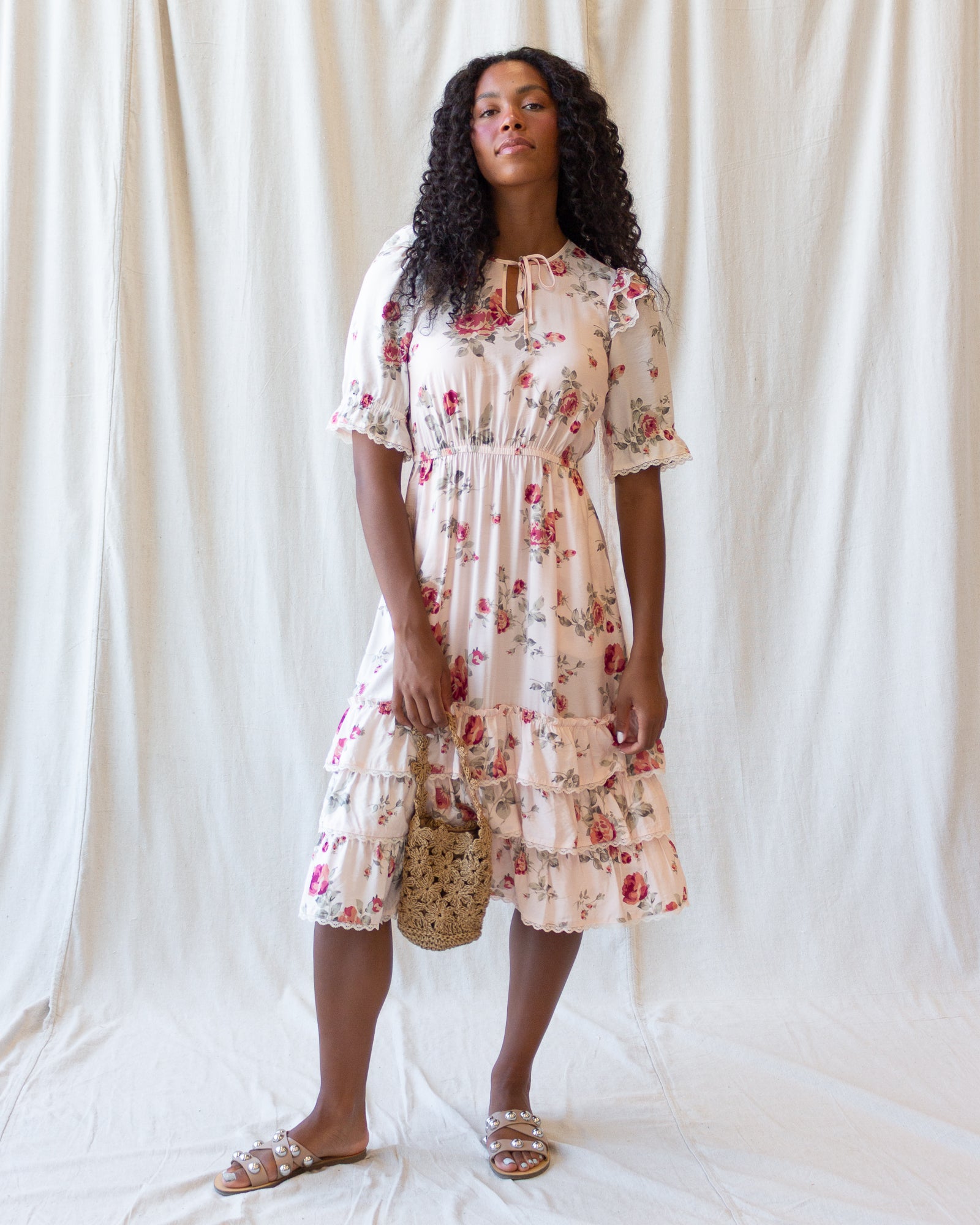 A woman stands against a draped white fabric backdrop wearing a short-sleeve cream dress with a red and pink floral print, holding a small woven bag and wearing flat sandals.
