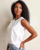 A woman with long, curly black hair wearing a white sleeveless top and light blue jeans, standing against a textured off-white wall.