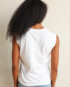 A woman with long, curly black hair wearing a white sleeveless top and light blue jeans, standing against a textured off-white wall.