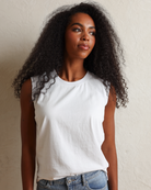 A woman with long, curly black hair wearing a white sleeveless top and light blue jeans, standing against a textured off-white wall.