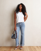 A woman with long, curly black hair wearing a white sleeveless top and light blue jeans, standing against a textured off-white wall.
