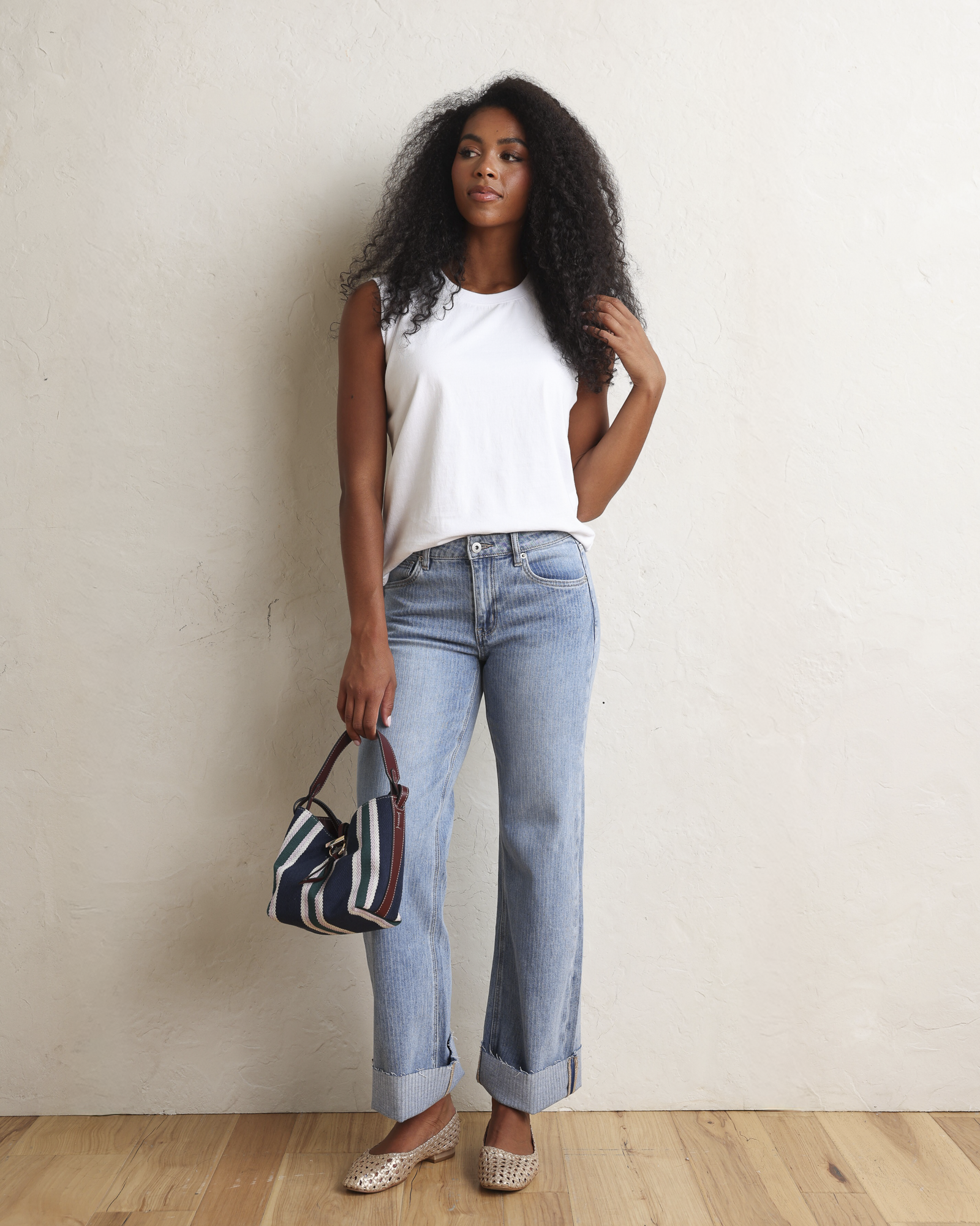 A woman with long, curly black hair wearing a white sleeveless top and light blue jeans, standing against a textured off-white wall.