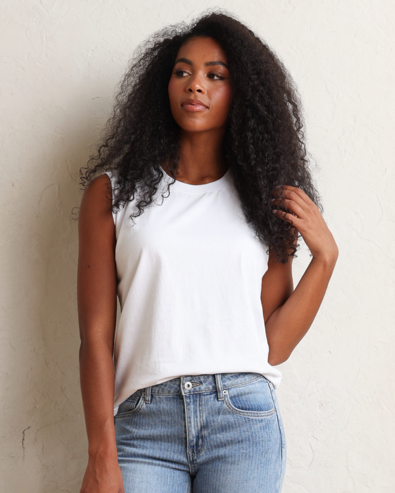A woman with long, curly black hair wearing a white sleeveless top and light blue jeans, standing against a textured off-white wall.