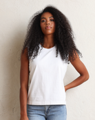 A woman with long, curly black hair wearing a white sleeveless tank top and light blue jeans, standing against a textured off-white wall.