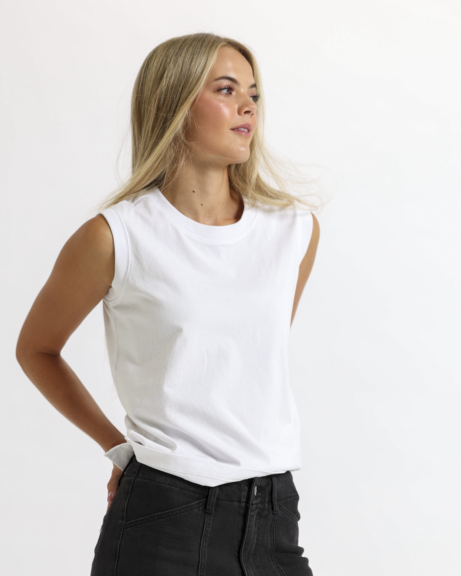 A woman with long, blonde hair wearing a white sleeveless top and black jeans, standing against a textured off-white wall.