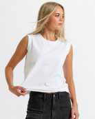 A woman with long, blonde hair wearing a white sleeveless top and black jeans, standing against a textured off-white wall.