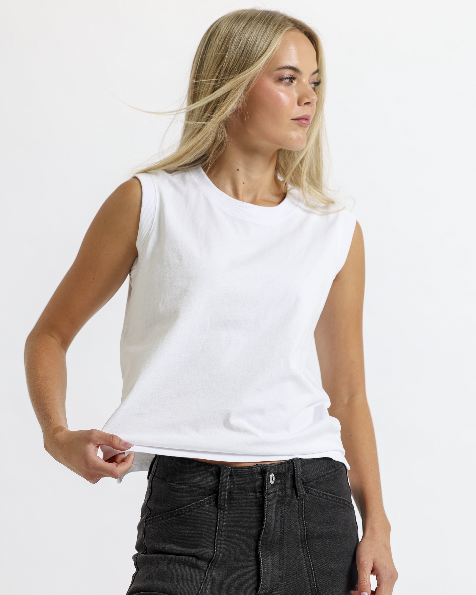 A woman with long, blonde hair wearing a white sleeveless top and black jeans, standing against a textured off-white wall.