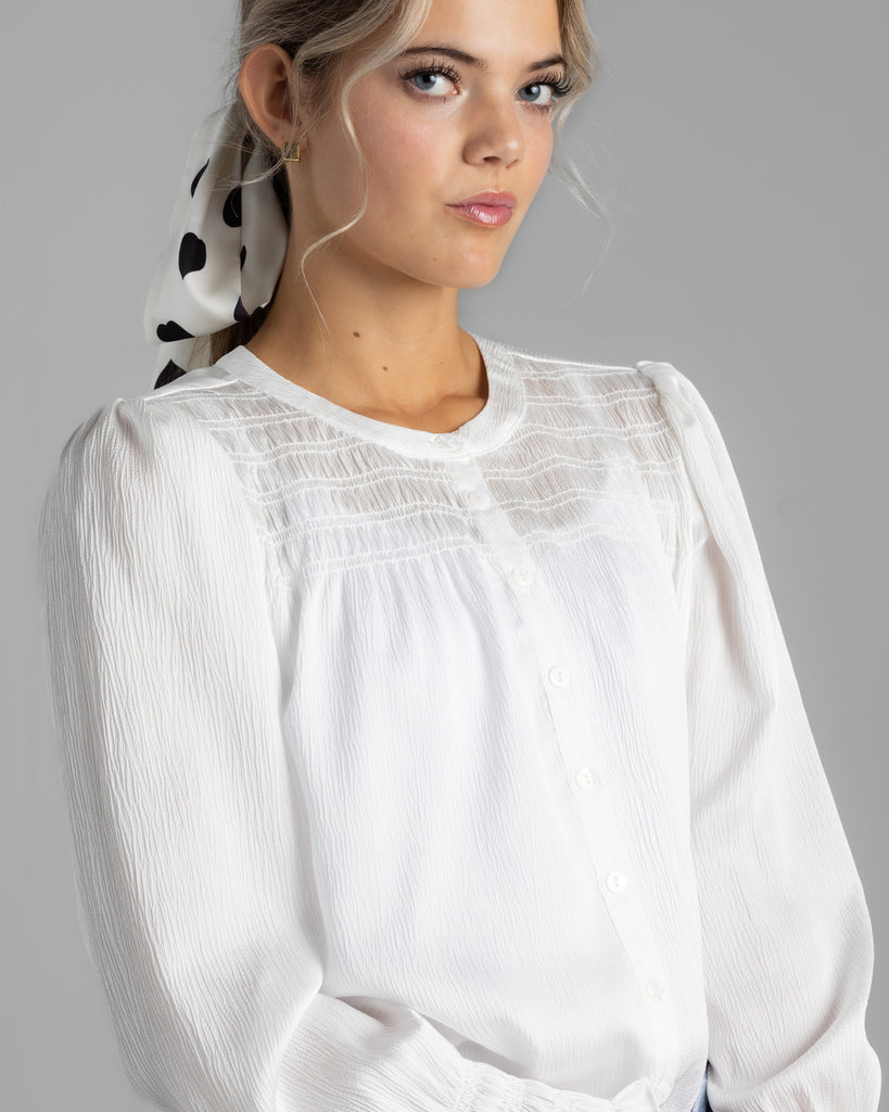 Model wearing a white long sleeve sheer blouse with smocking on yoke.