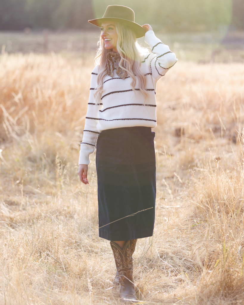 Woman wearing a cowboy hat, striped sweater, skirt, and cowboy boots outside.