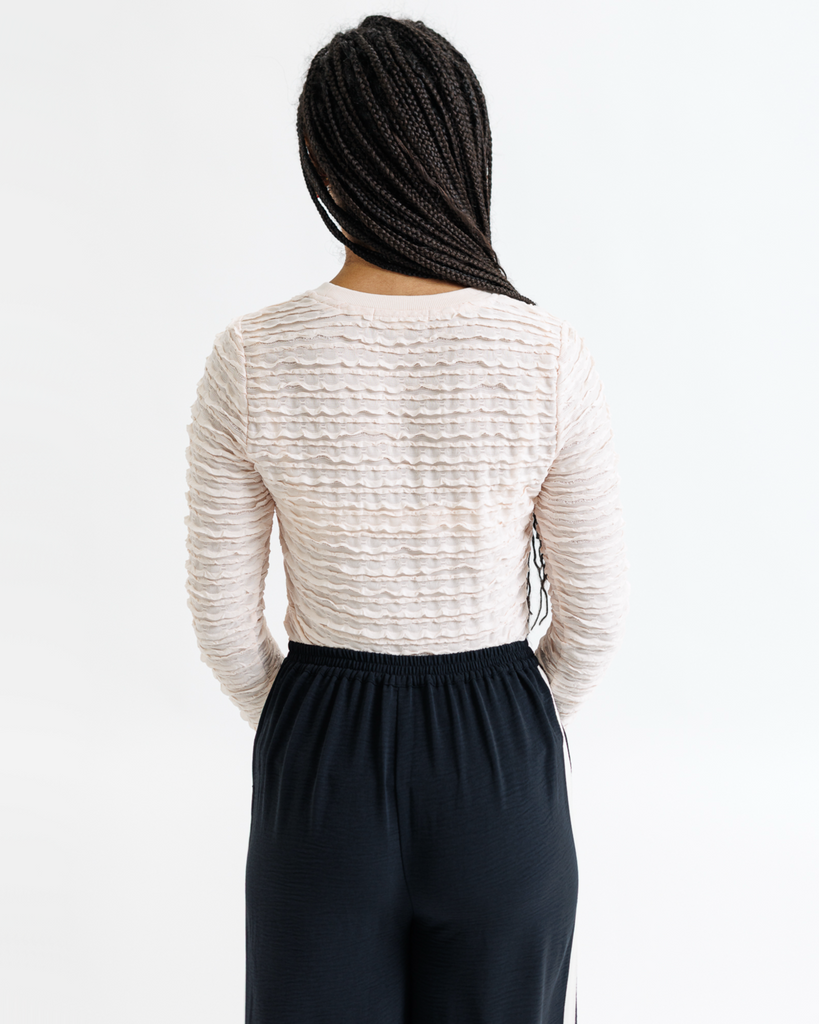 Woman wearing a textured cream long-sleeve top and dark pants on a white background