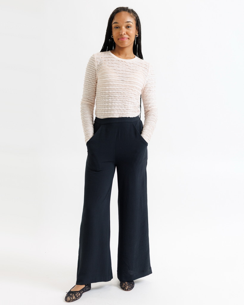 Woman wearing a cream lace top and dark wide-leg pants on a white background