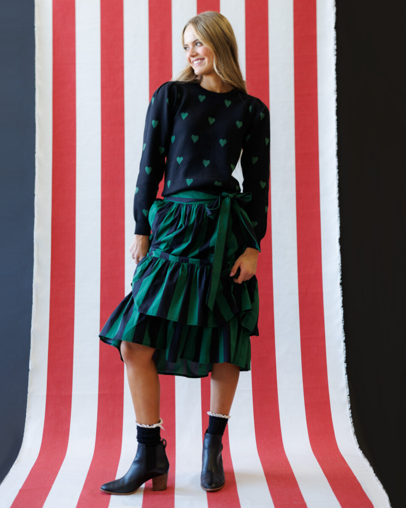 Woman wearing a black sweater with green heart patterns and a green skirt against a striped background