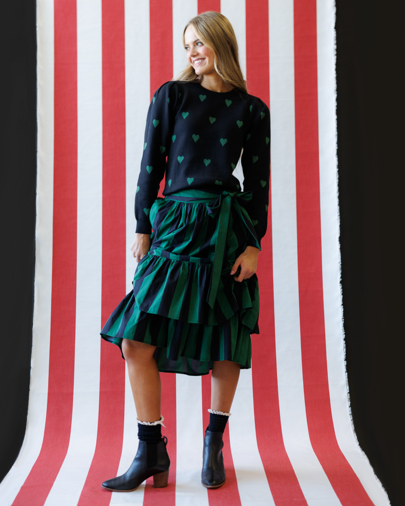 Woman wearing a black sweater with green heart patterns and a green skirt against a striped background