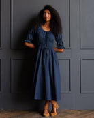 A woman with curly black hair wearing a navy blue midi dress with puff sleeves and a drawstring waist, standing against a dark gray paneled wall.