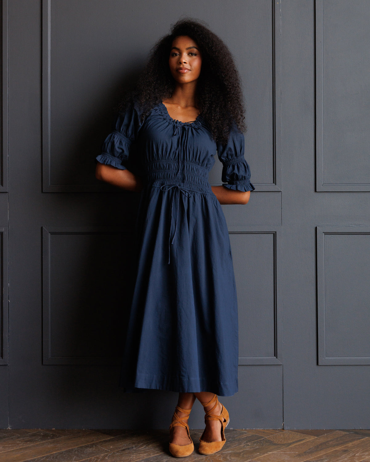 A woman with curly black hair wearing a navy blue midi dress with puff sleeves and a drawstring waist, standing against a dark gray paneled wall.