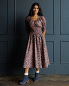 A woman stands against a dark paneled wall wearing a dusty rose midi dress with a pink floral pattern, puff sleeves, and a smocked waist. She is wearing blue suede ankle boots with white socks.