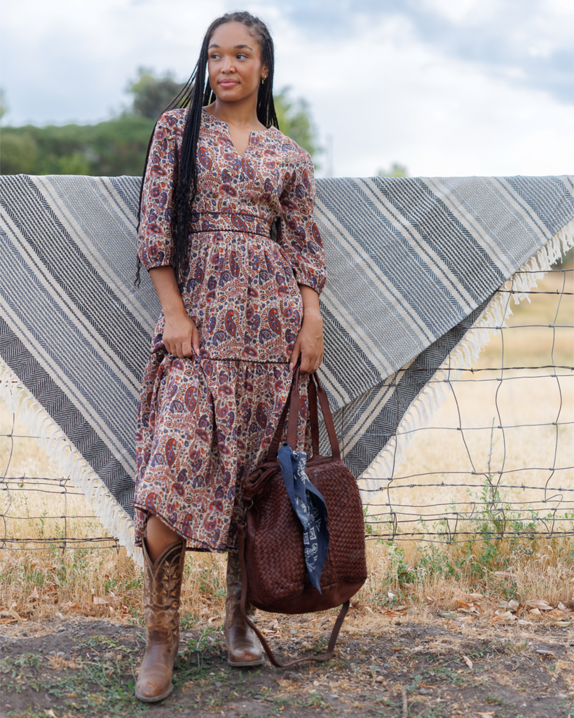 Woman holding a brown purse and wearing a multicolored dress and brown cowboy boots standing next to a fence.