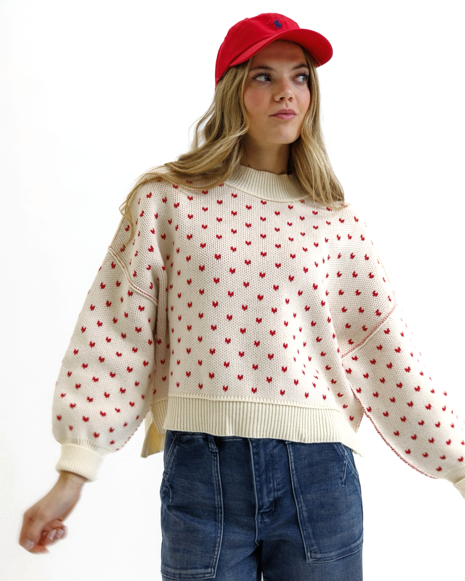 Woman wearing a cream sweater with red heart patterns, blue jeans, and a red cap on a white background