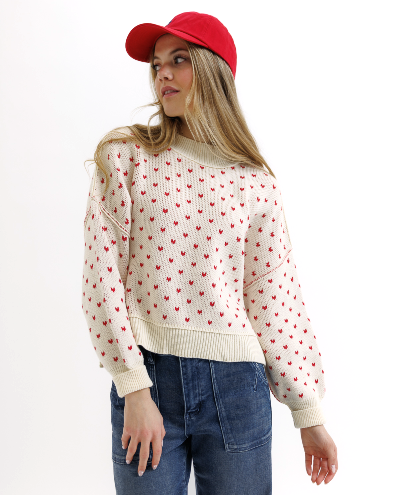 Woman wearing a cream sweater with red heart patterns, blue jeans, and a red cap on a white background