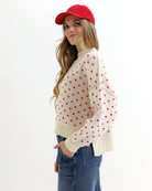 Woman wearing a cream sweater with red heart patterns, blue jeans, and a red cap on a white background