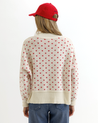 Woman wearing a cream sweater with red heart patterns, blue jeans, and a red cap on a white background