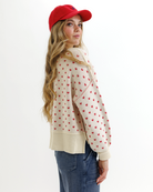 Woman wearing a cream sweater with red heart patterns, blue jeans, and a red cap on a white background