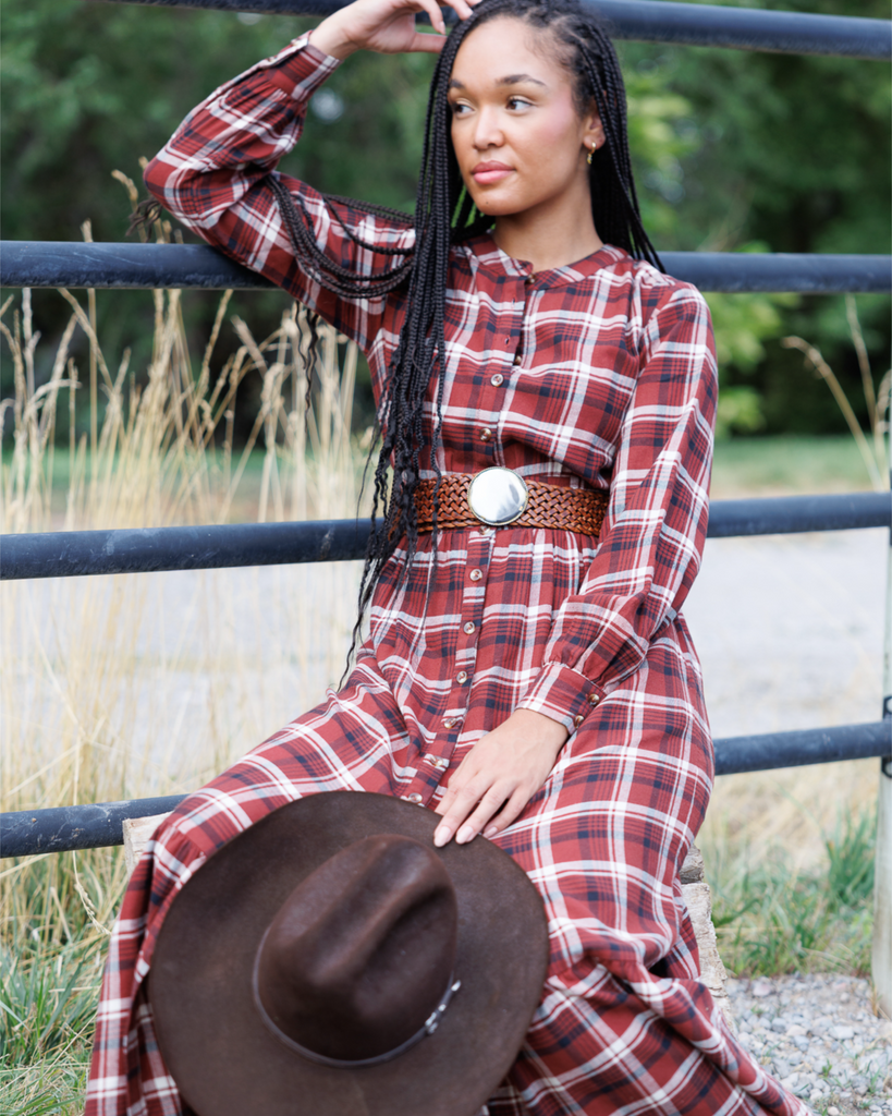 Woman in a red plaid dress with a cowboy hat sitting on a fence outdoors.