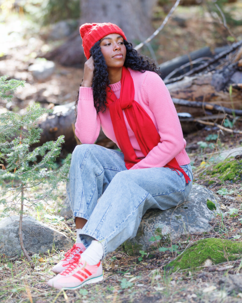 Woman wearing a pink sweater, red beanie, and scarf sitting in a forest.