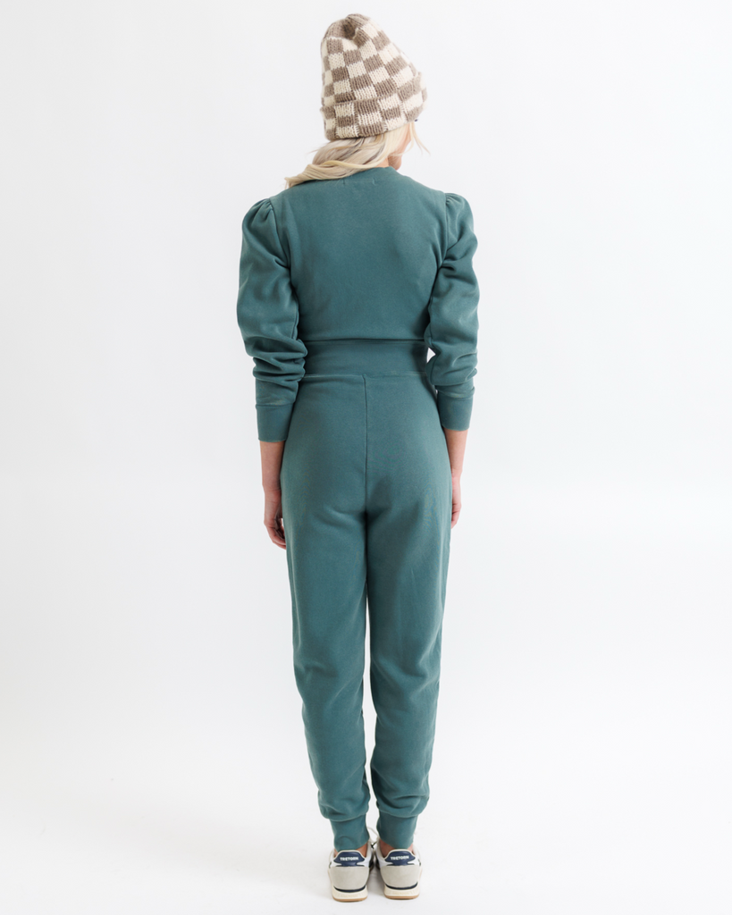 Woman wearing a teal jumpsuit with a checkered beanie on a white background
