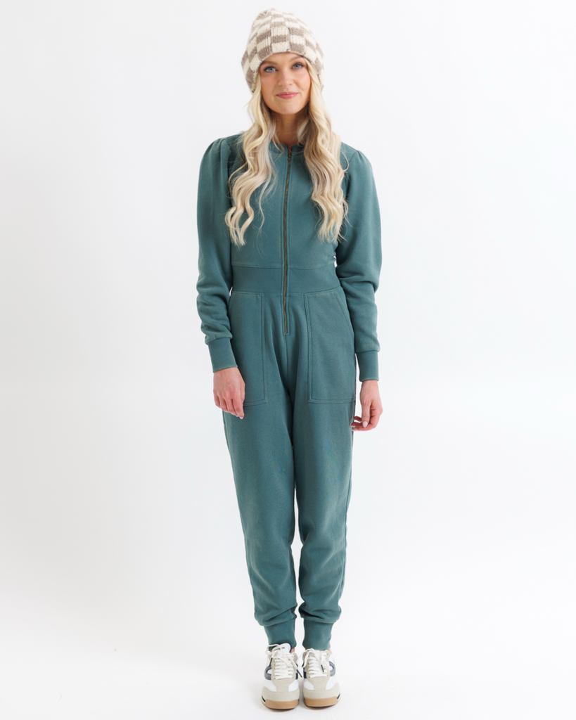 Woman wearing a teal jumpsuit with a checkered beanie on a white background