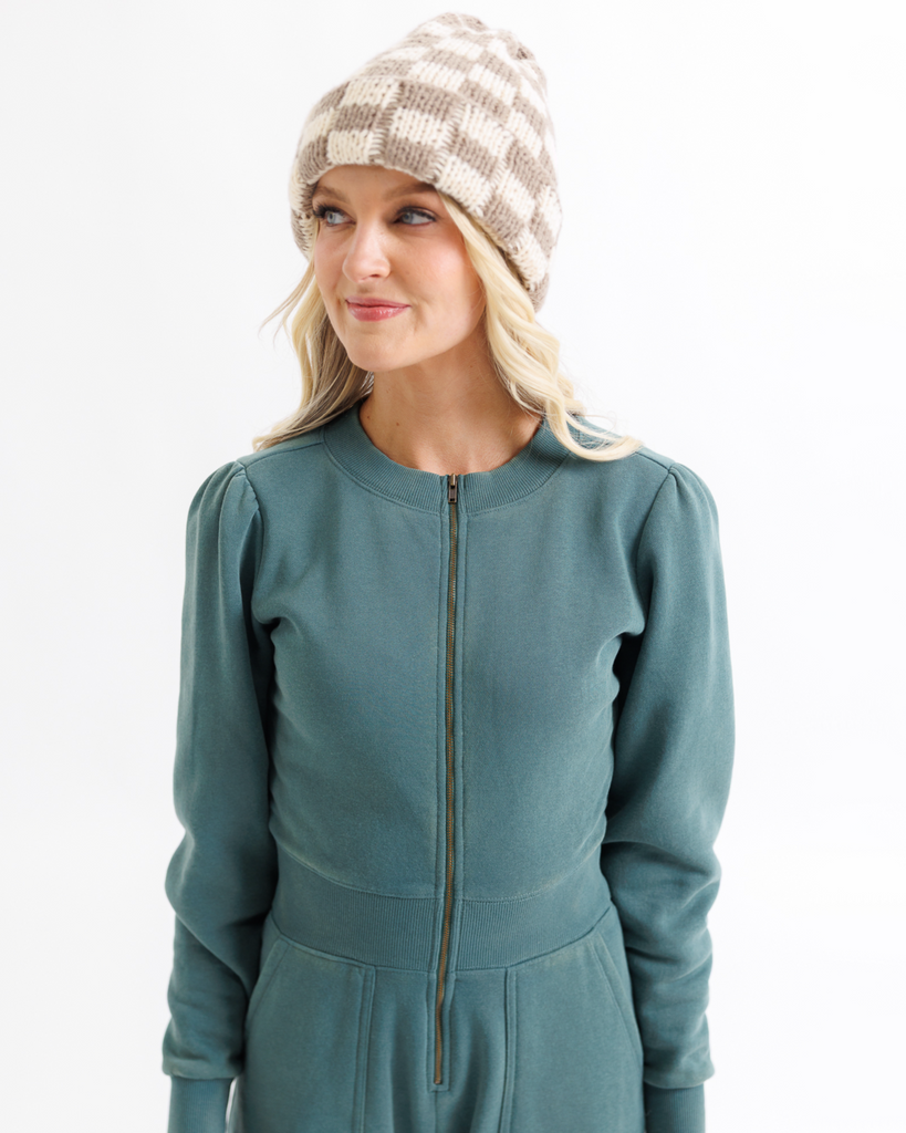 Woman wearing a teal jumpsuit and checkered beanie on a white background