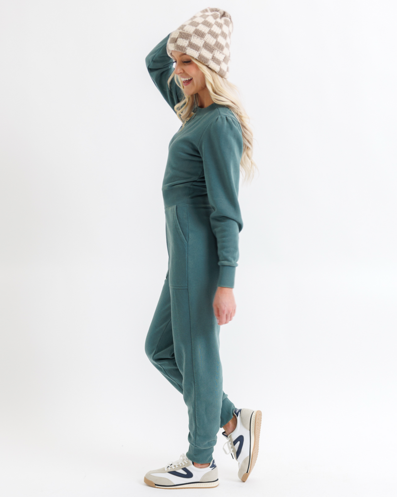 Woman wearing a teal jumpsuit with a checkered beanie on a white background